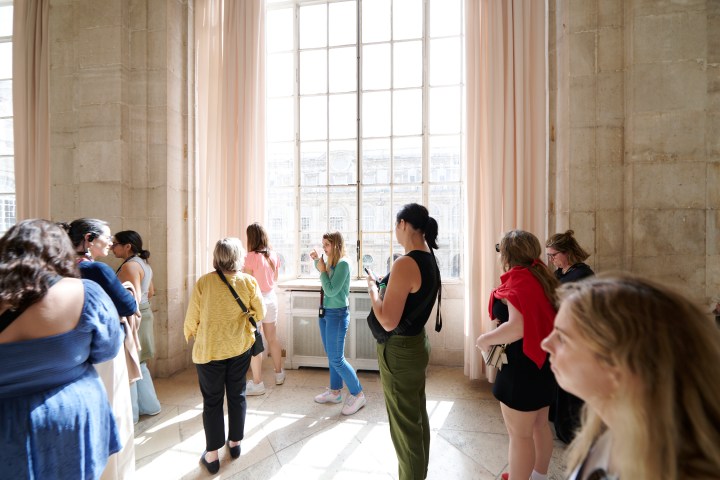 People standing in a sunlit room with large windows and curtains.