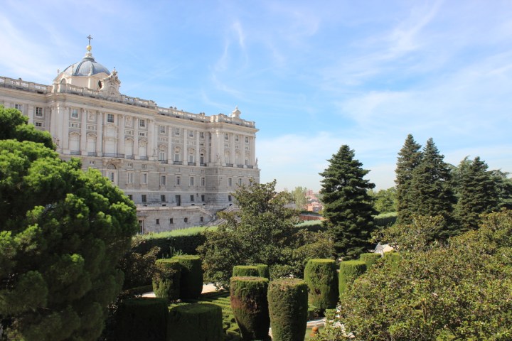 Elegant palace with intricate facade beside manicured gardens and trees under a clear blue sky.