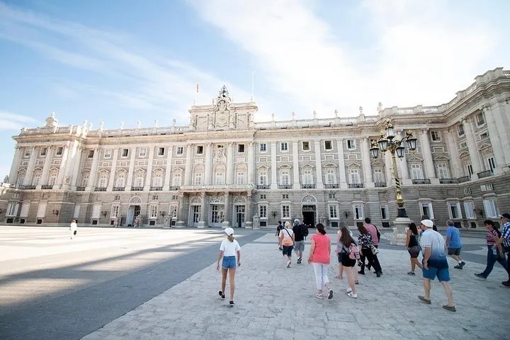 Tourists walk towards a large, ornate palace under a bright blue sky.