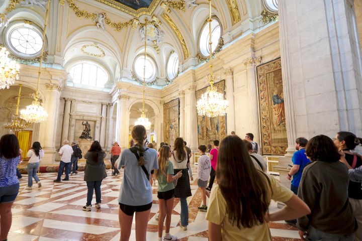 Tour group in ornate palace room with high ceilings and chandeliers.
