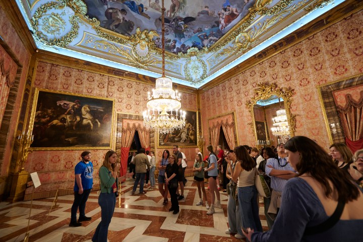 Group of people touring an ornate, decorated room with chandeliers and paintings.