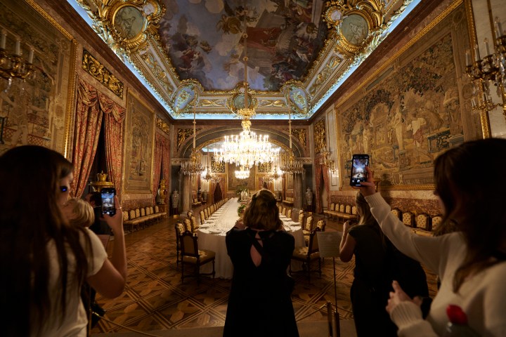 People photographing an ornate dining room with chandeliers and tapestries.