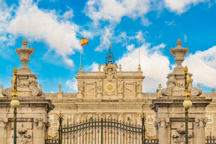 Grand building with ornate gates and a flag atop, against a cloudy blue sky.